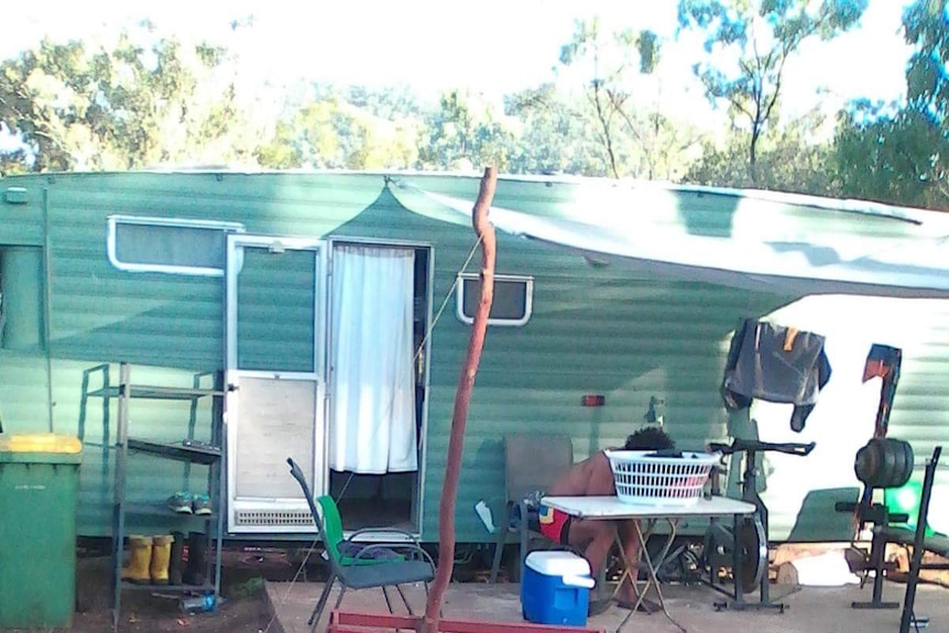 Green caravan, stick holding shade cloth, man seen hiding face at an outdoor table, rough conditions