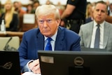 Donald Trump sits at a desk in court, wearing a blue suit and tie and looking unsatisfied.