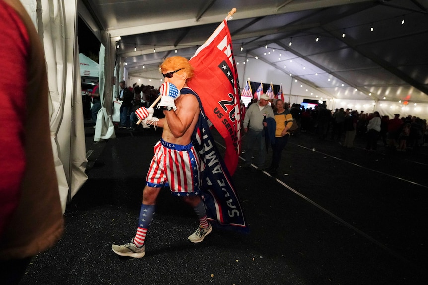 man in a trump wog and us flag boxers walks with a giant red herschel flag