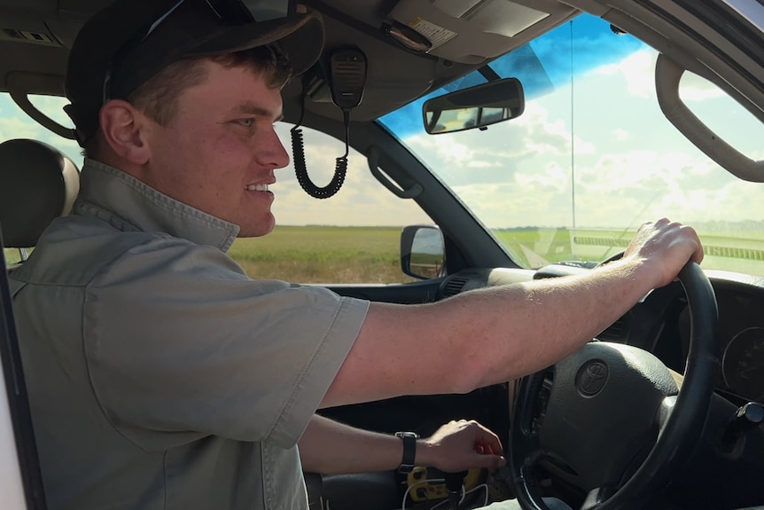 A young man wearing a khaki shirt and a black cap is driving his ute.