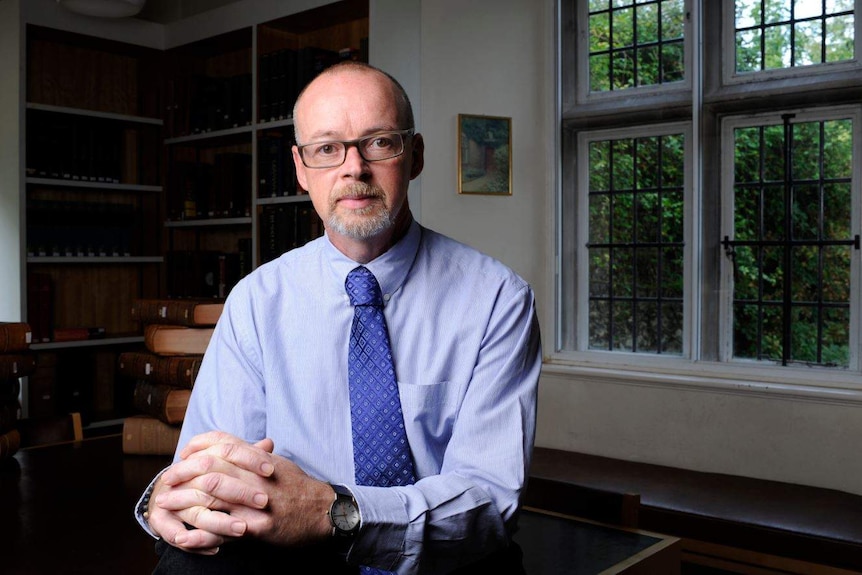 A man with a neat beard and glasses, wearing a shirt with tie, sits at a desk with hands folded. Bookshelves are in background.