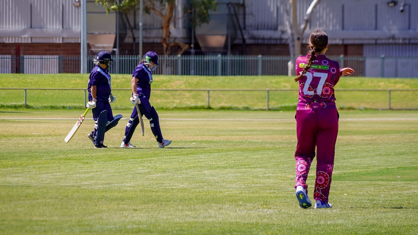 Three female cricketers, two batters and a bowler walk onto a grass oval