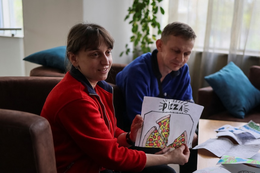 A woman and a man sit at a coffee table with the woman holding a children's draw of a pizza.