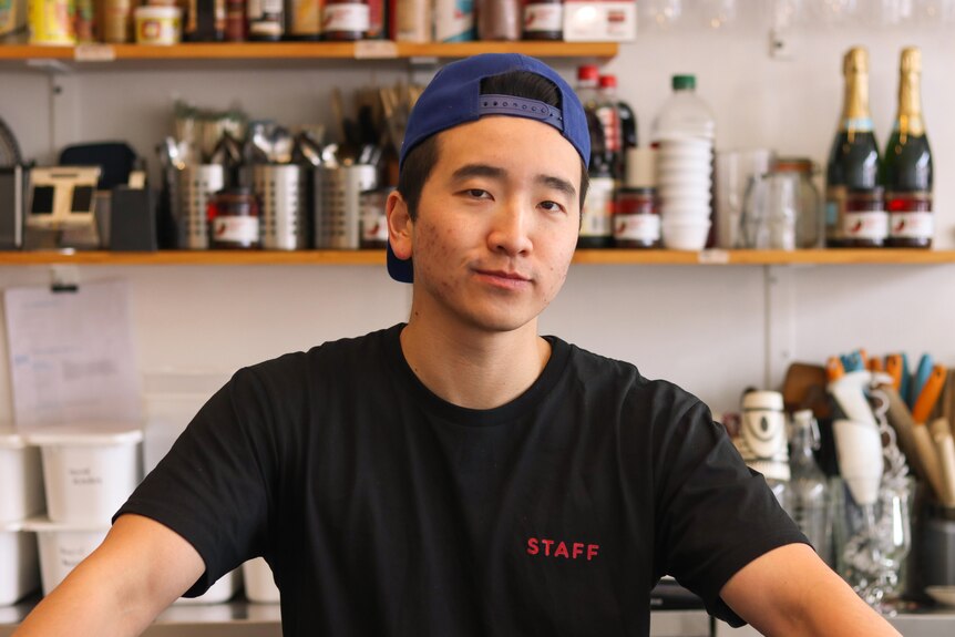 Ethan Yong wears a black t-shirt and stands in front of shelves in a kitchen