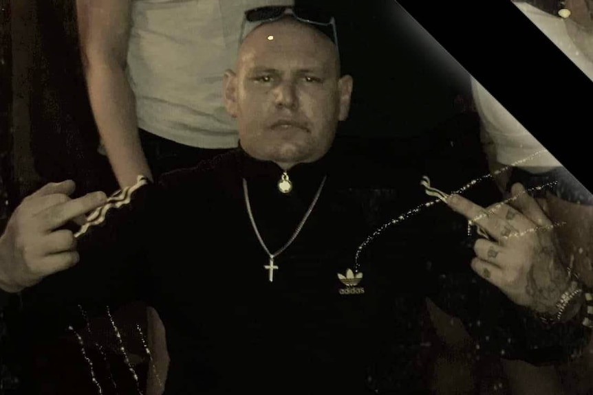 A with a serious expression wears a dark tracksuit, a chain around his neck, and sunglasses on his head.