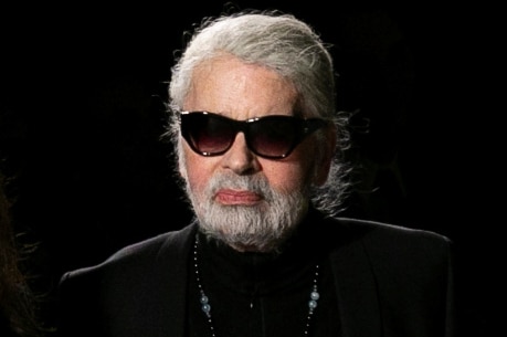 A bearded Karl Lagerfeld, wearing dark glasses and ornate jewellery, stands against a black background.