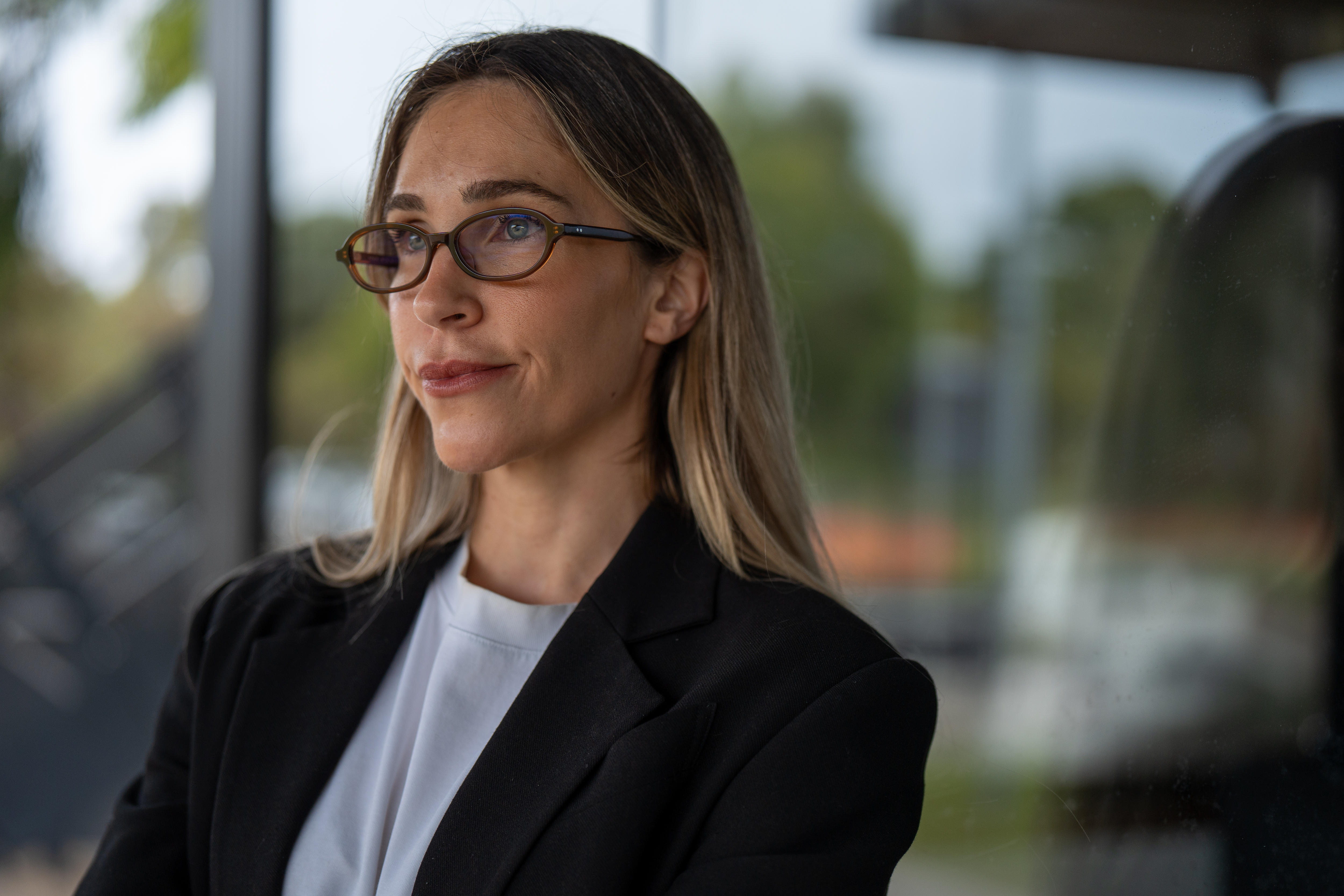 A woman with glasses looks away from the camera while standing next to a glass window