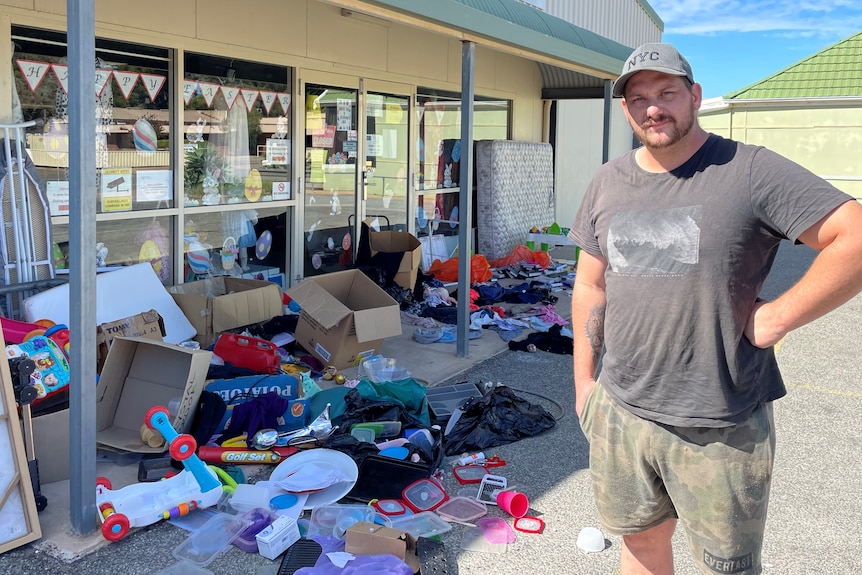 Man with hand on hip stands in front of a shop with piles of clothes strewn across the footpath.