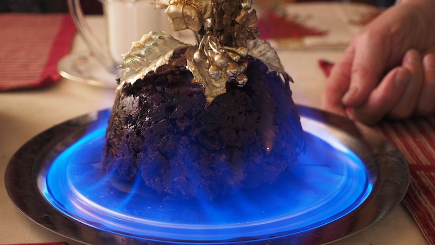 A traditional plum pudding on a plate, surrounded by a ring of blue flame, and topped with gold decorations.