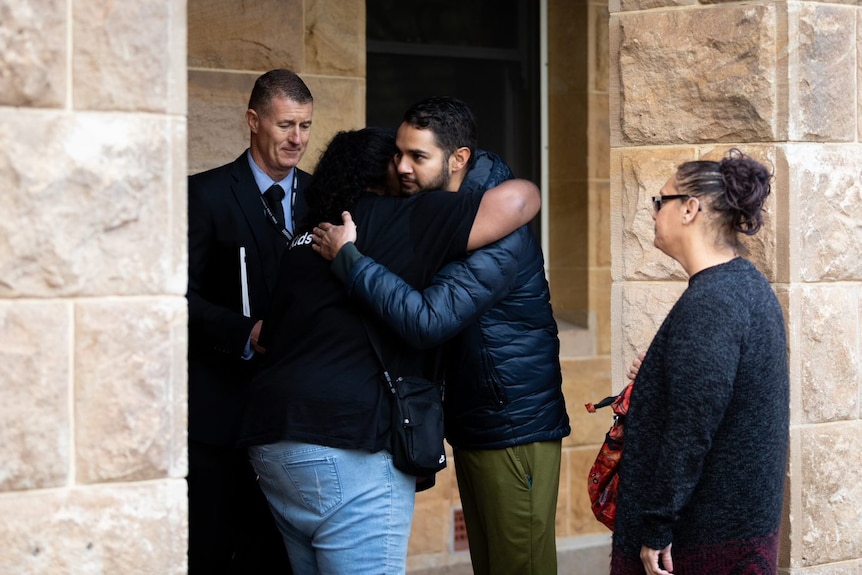 A picture of an Indigenous man and woman hugging outside court as a detective looks at them and another woman looks on.
