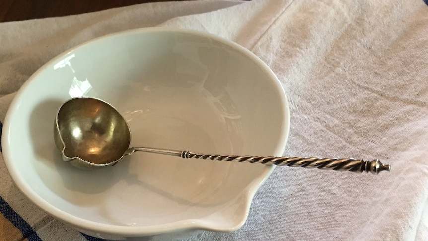 A silver ladle with a twisted handle sits in a white bowl on a table covered by a cloth.