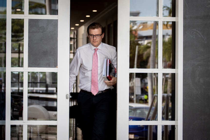 A man in a white shirt and pink tie exits a courthouse through sliding doors. 