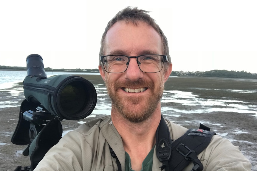 A man with glasses and short brown hair takes a selfie on a beach with a large black camera lens behind him.