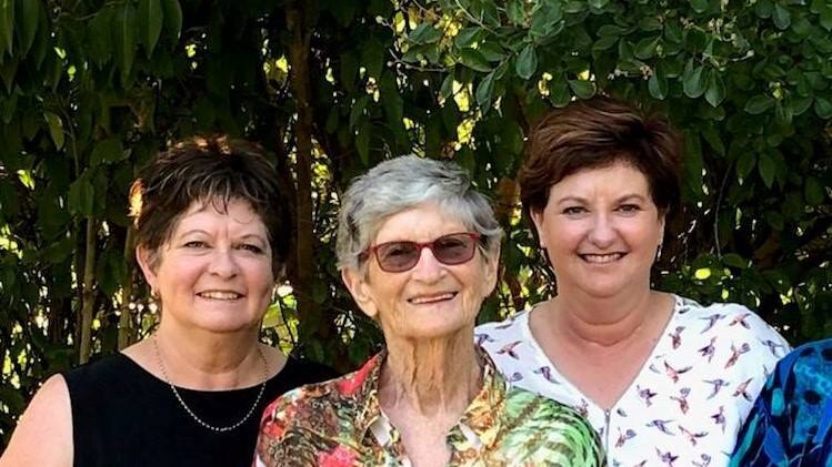 Three women stand smiling towards the camera in front of shrubbery.