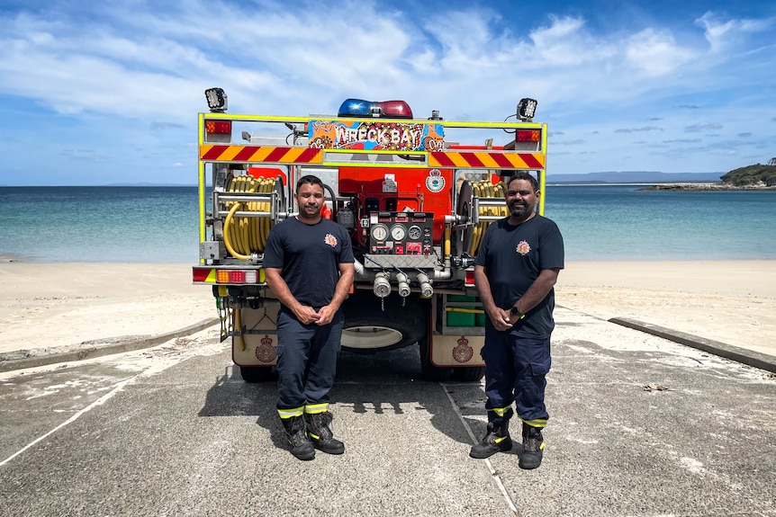 Two firefighters stand on the beach in front of a fire truck with decals.