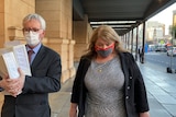 A woman in a grey dress and black blazer walks beside a man in a suit, holding thick folders, outside a court building