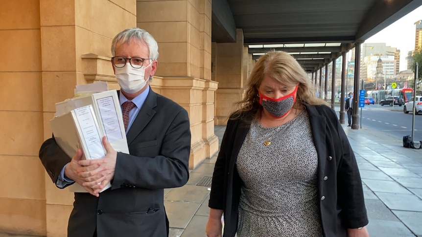 A woman in a grey dress and black blazer walks beside a man in a suit, holding thick folders, outside a court building