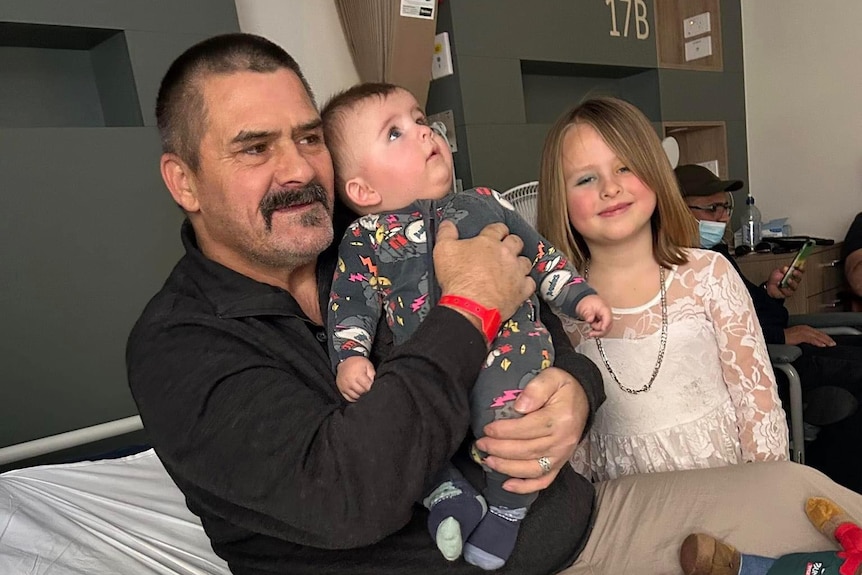 A man with a moustache holding a small baby, with a young girl standing next to them.