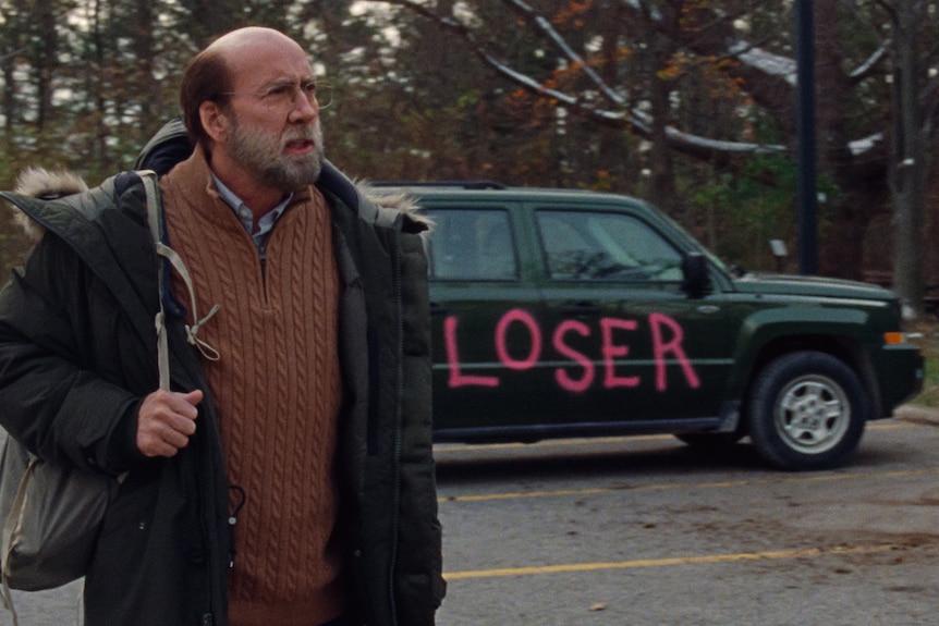 Nicolas Cage in character as an academic, carries a bag over his shoulder in a carpark, a vehicle behind him spraypainted loser.