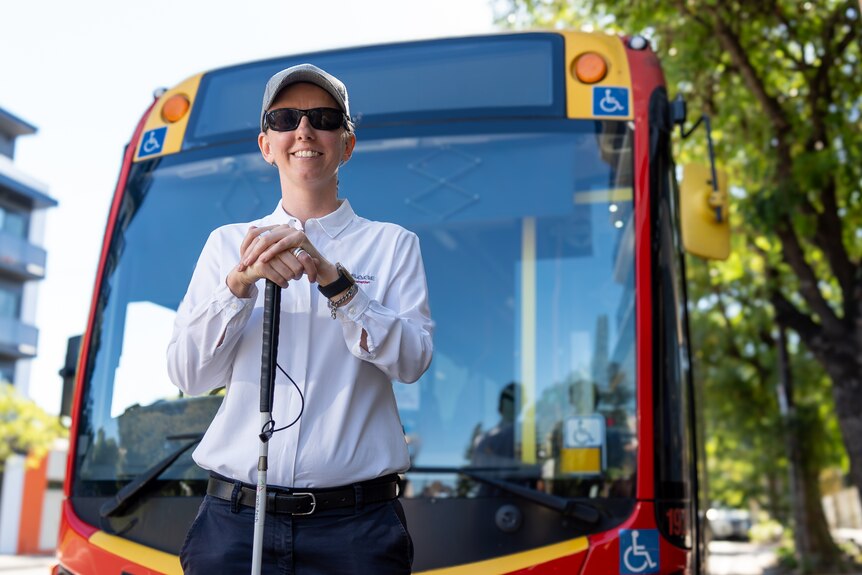 A woman wearing a cap, sunglasses and holding onto a walking stick stands in front of a public bus.
