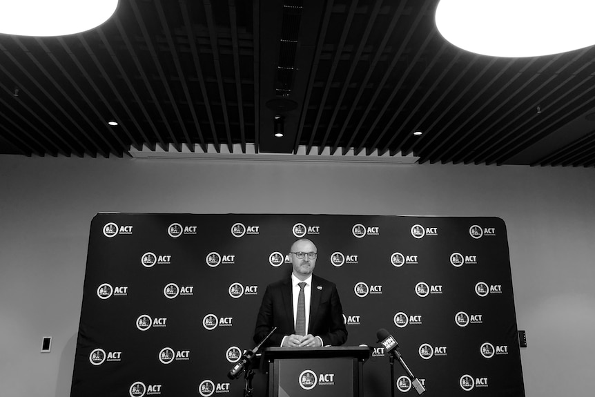 A black and white photo of Andrew Barr standing at a lectern with bright lights shining on him
