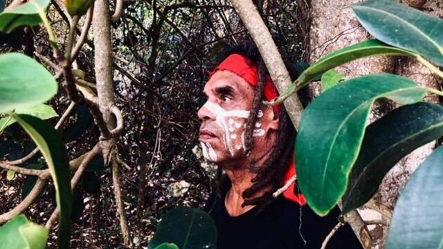 Side portrait of Gumbaynggirr man with red bandana and white face paint wearing a black shirt in the bush
