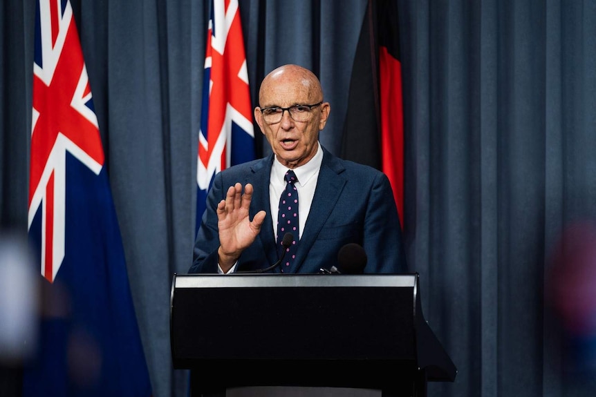 John Quigley standing at a podium for a press conference, in front of Australian flags and a blue curtain.