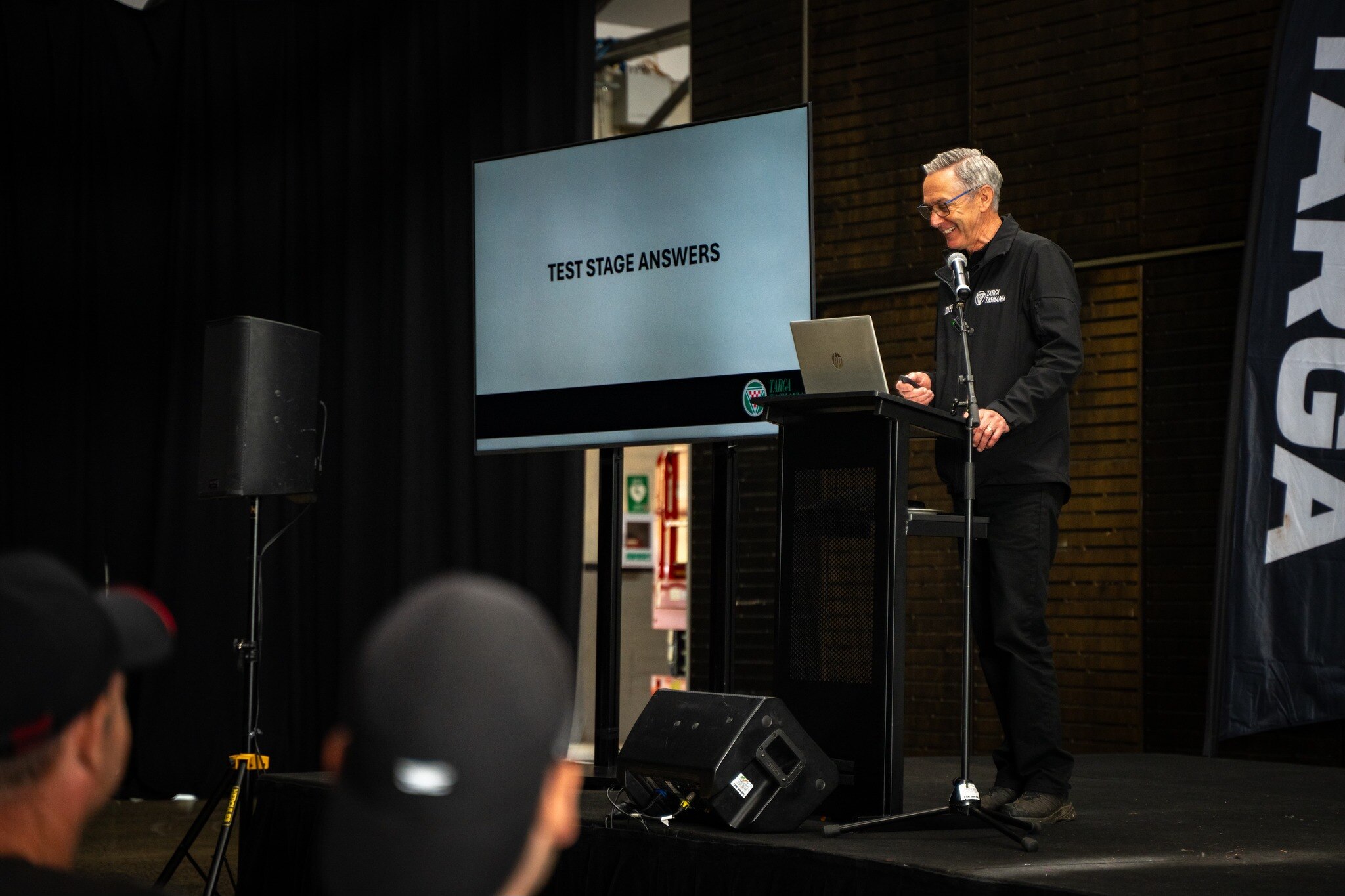 A man stands on a stage giving a presentation with a screen that says 'test stage answers'.