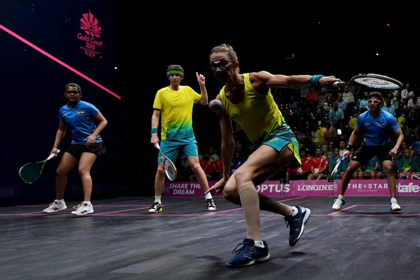A female squash player in a yellow shirt and gold shorts plays a shot in front of three other players.