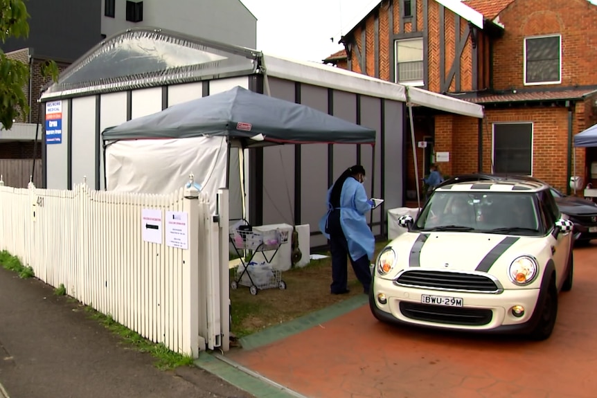 A white car in a driveway of a home next to a white covid-testing tent. A woman in blue PPE walks with a clipboard