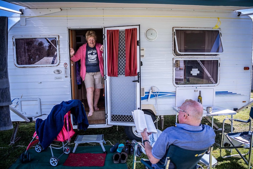 An older woman wearing a pink jumper walking out of a caravan and a man reading a book.