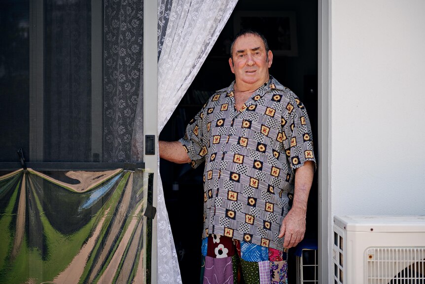 An older man stands in a doorway, holding back a lace curtain