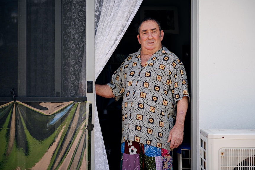 An older man stands in a doorway, holding back a lace curtain