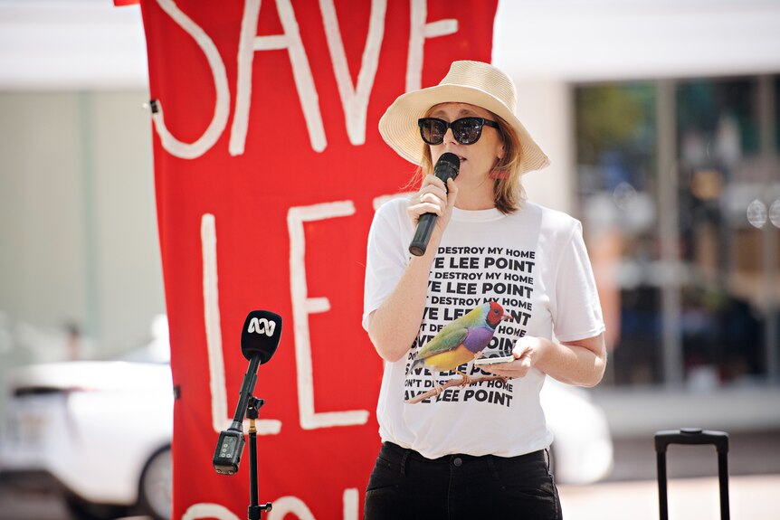 A woman in a brimmed hat and sunglasses speaking in front of a sign, at a protest.