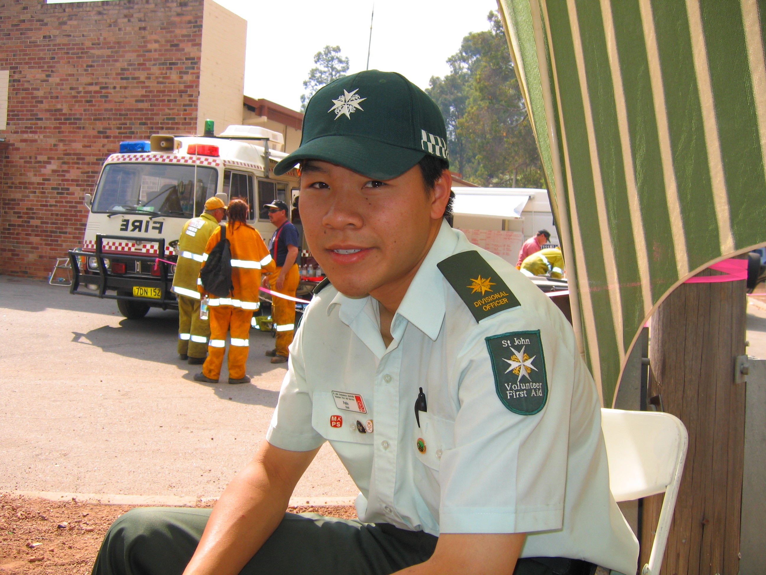 Um adolescente de Hong Kong, vestido com boné e camisa verde da St John Ambulance, com distintivo, veículo de emergência atrás dele.