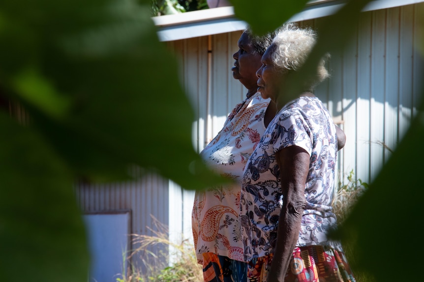 Two women are outside a large building. They are being seen through the trees.