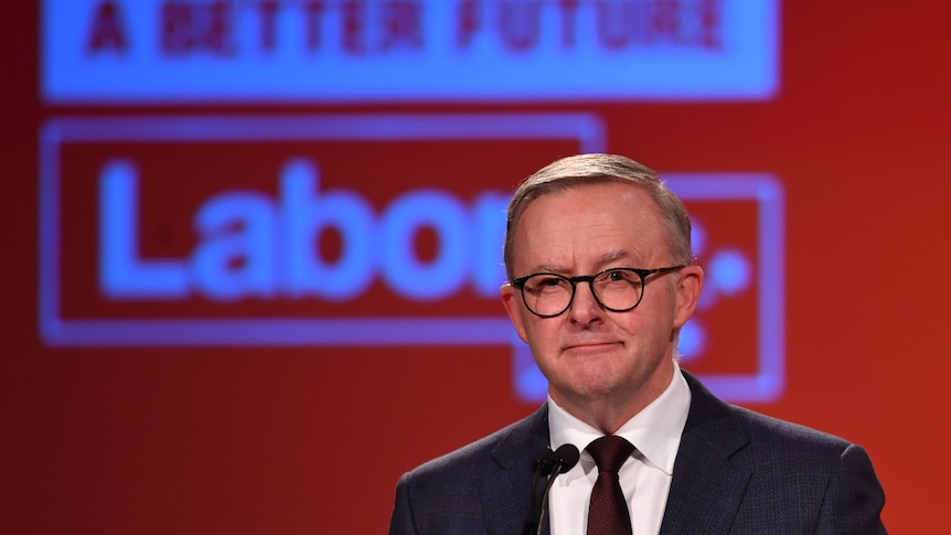 Anthony Albanese in a black suit and tie wearning glasses stands at a microphone in front of a red background with a Labor logo