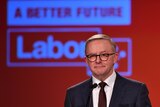 Anthony Albanese in a black suit and tie wearning glasses stands at a microphone in front of a red background with a Labor logo