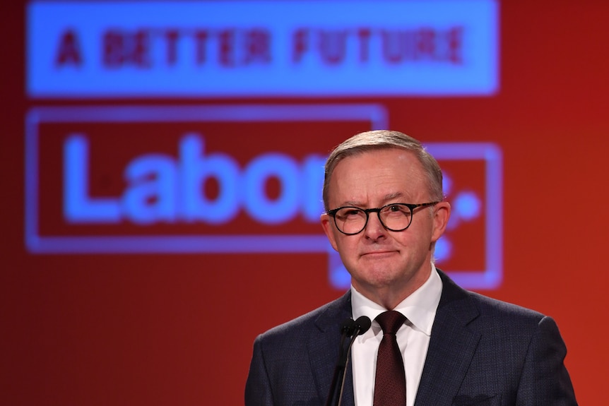 Anthony Albanese in a black suit and tie wearning glasses stands at a microphone in front of a red background with a Labor logo