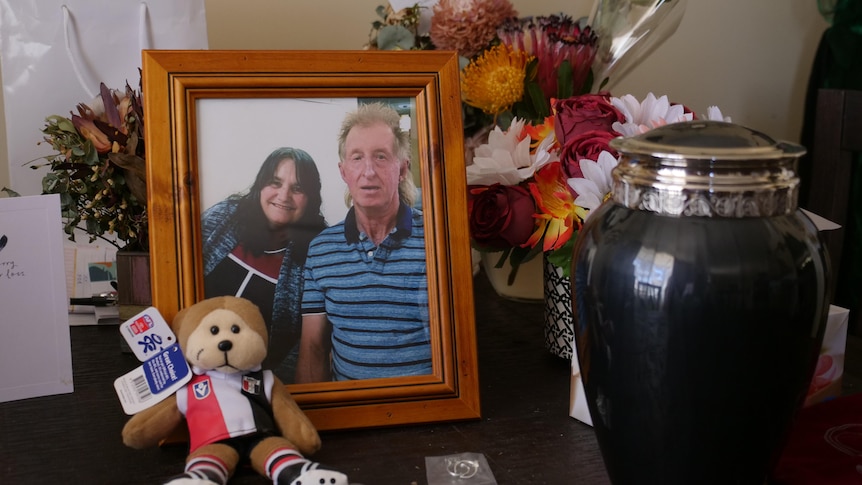 On a table, a photo of a man and a woman happy to be together, an urn, flowers and a plush bear wearing the St Kilda Saints kit.