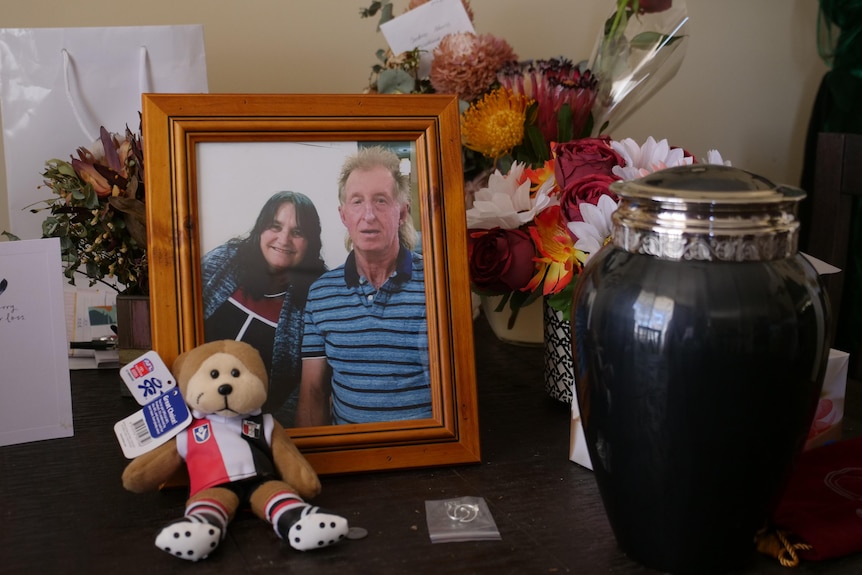 On a table, a photo of a man and a woman happy to be together, an urn, flowers and a plush bear wearing the St Kilda Saints kit.