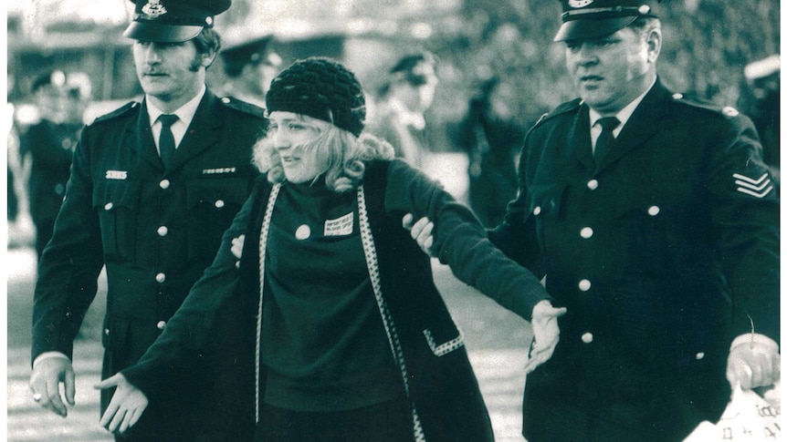 black and white photo of woman in coat and cap being held on both sides by police officers