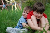 A boy cuddles his big brother as they sit in the grass smiling at the camera