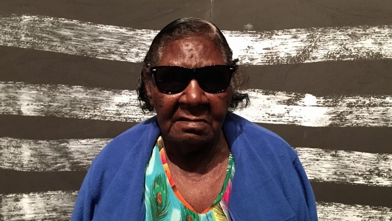 An old woman wearing sunglasses, a blue jumper and a floral top stands in front of a painting of black and white stripes.