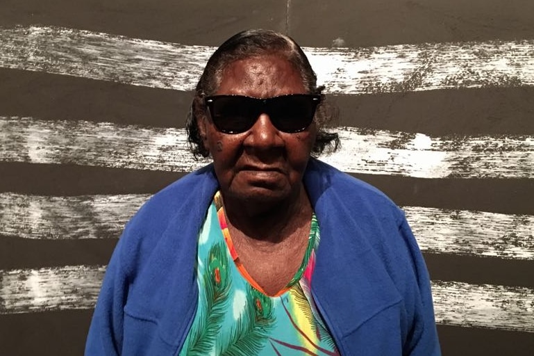 An old woman wearing sunglasses, a blue jumper and a floral top stands in front of a painting of black and white stripes.