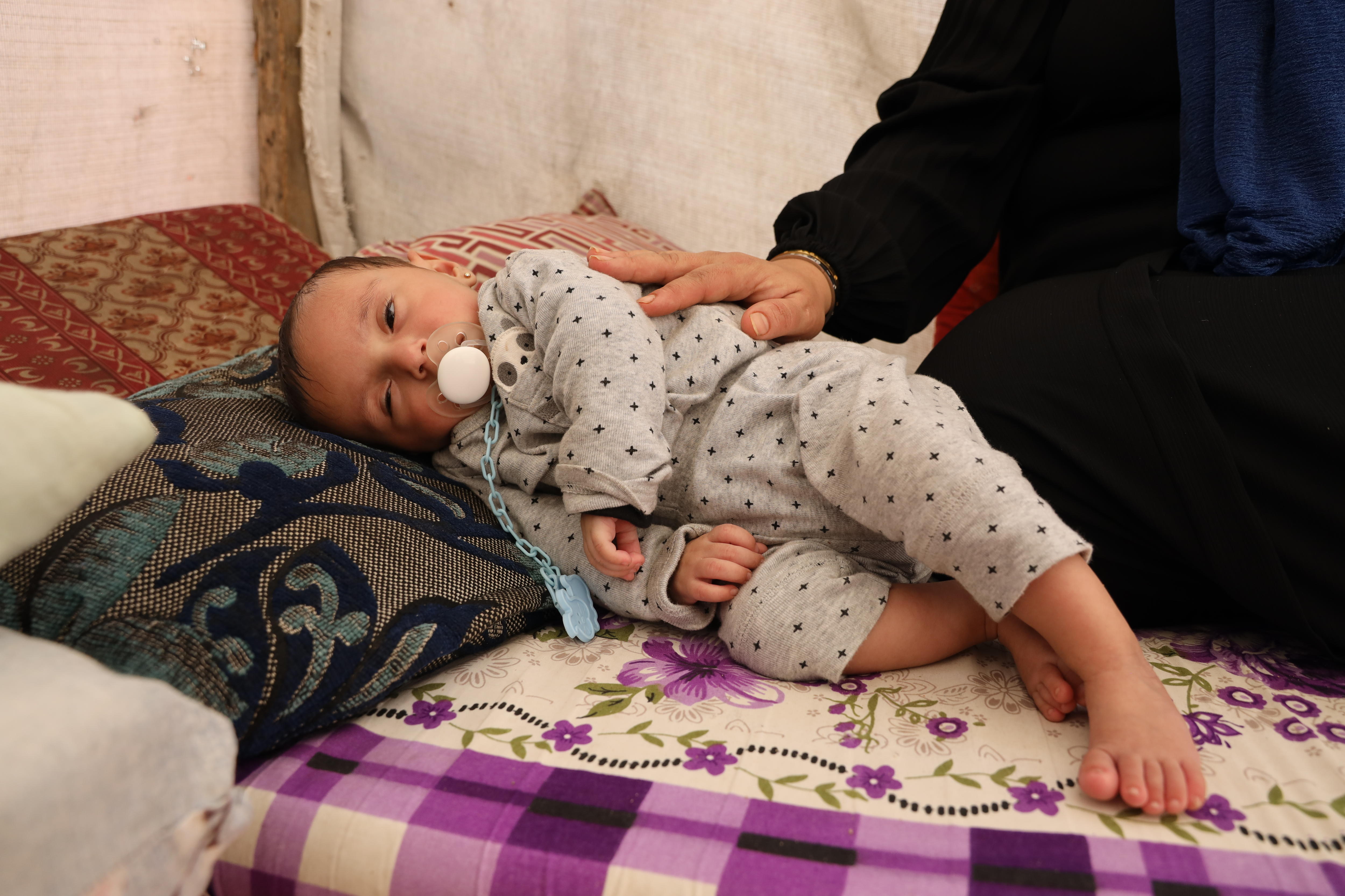 A seven-month-old baby looks sleep as she lays on her side, the comforting hand of her mother on her back.