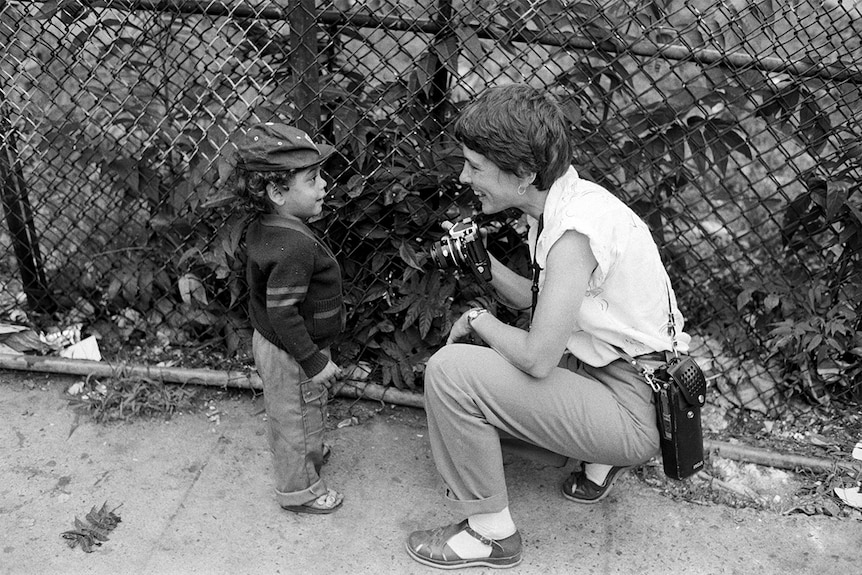 A young child with cap and cardigan speaks to crouched down woman holding camera in front of wire fencing and trees.