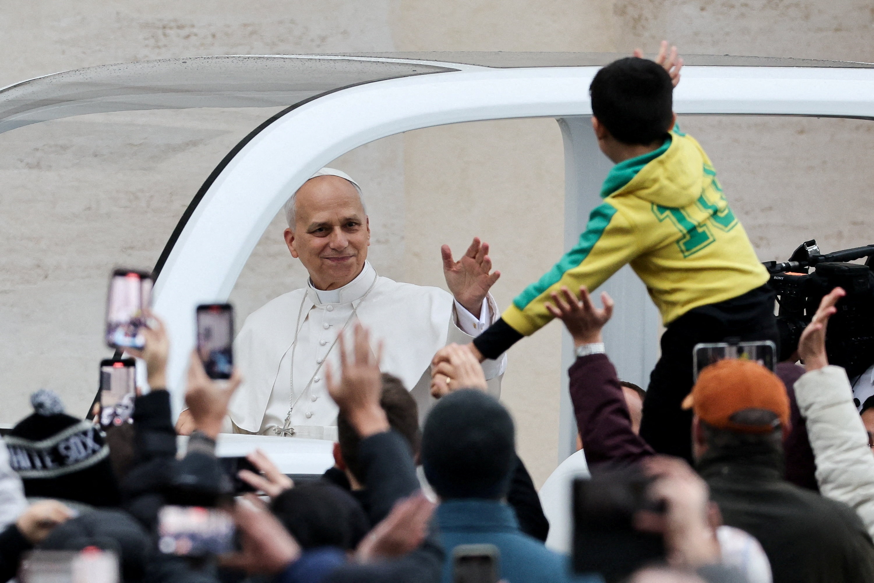 Pope Leo in the popemobile waving while people take photos on phones.
