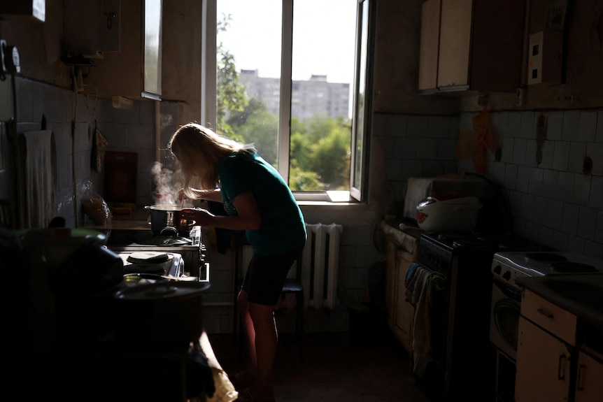 A woman cooks in the kitchen in front of a window showing trees and buildings outside. 