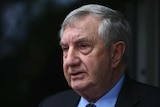 Profile of an older man with greying hair in a suit speaking to journalists